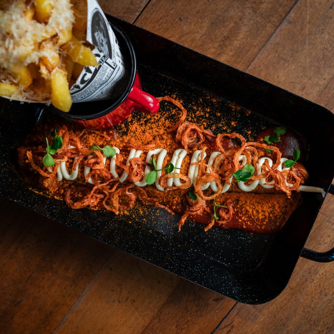 Gebackene Speisen im schwarzen Bratdeckel: cremige Tomatensauce, Zwiebelringe, pürierte Kräuter, Weißkohl-Dekoration, knusprige Garnelen, klecks Sahne, daneben frittierte Kartoffeln im Papiergefäß.